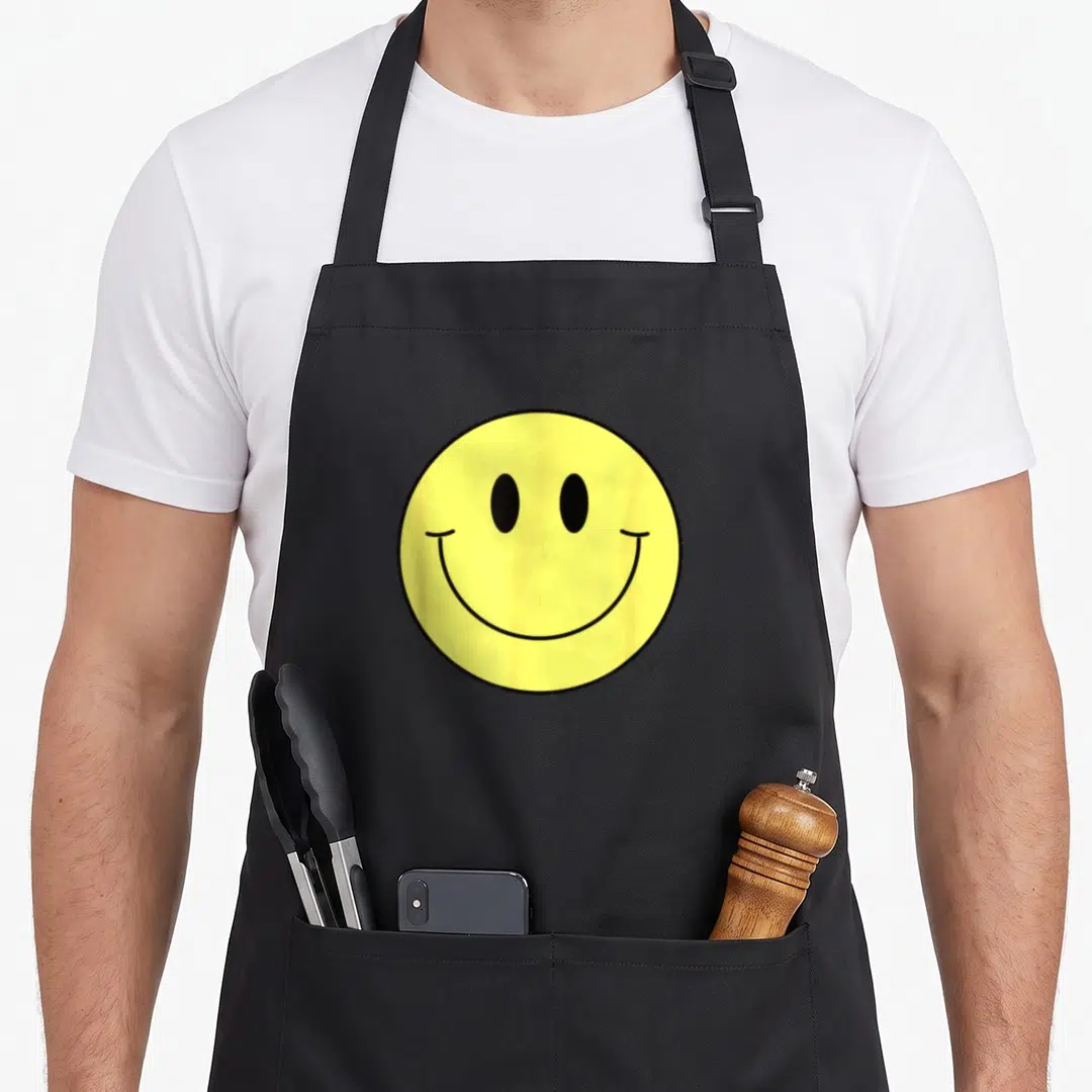 70s face on apron - Yellow smiley face with black eyes and mouth on black background — shown on a black cotton canvas apron against a white background