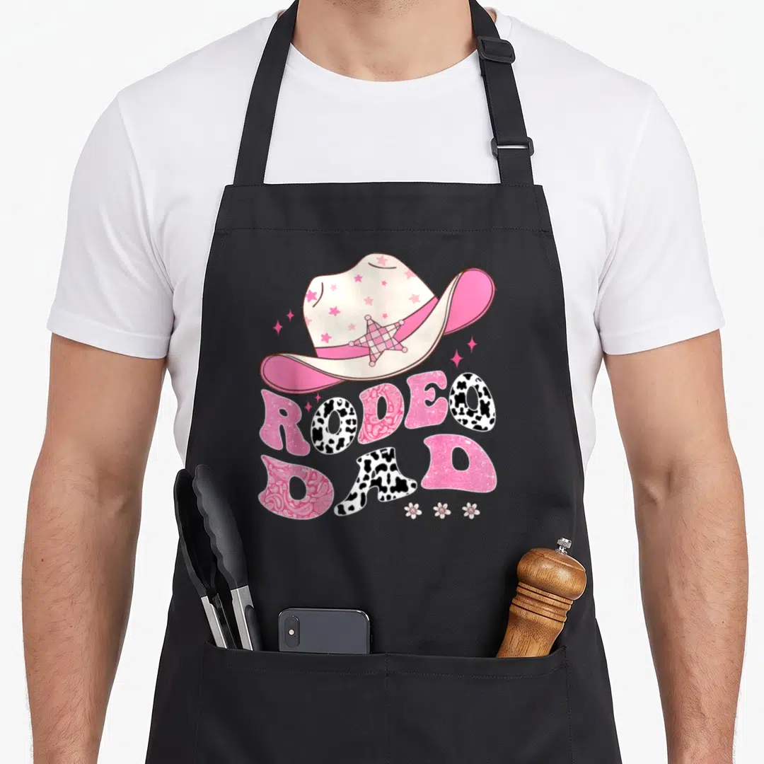1st cowgirl aprons - Pink cowboy hat with stars above RODEO DAD text in cow print and paisley patterns with three — shown on a black cotton canvas apron against a white background