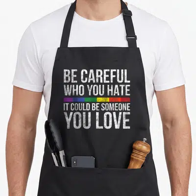 Careful black pride apron - White text BE CAREFUL WHO YOU HATE IT COULD BE SOMEONE YOU LOVE with a multicolored rainbow — shown on a black cotton canvas apron against a white background