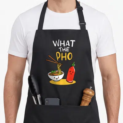 Noodle pho apron - Cartoon bowl of pho with chopsticks and smiling sriracha bottle under stacked text — shown on a black cotton canvas apron against a white background