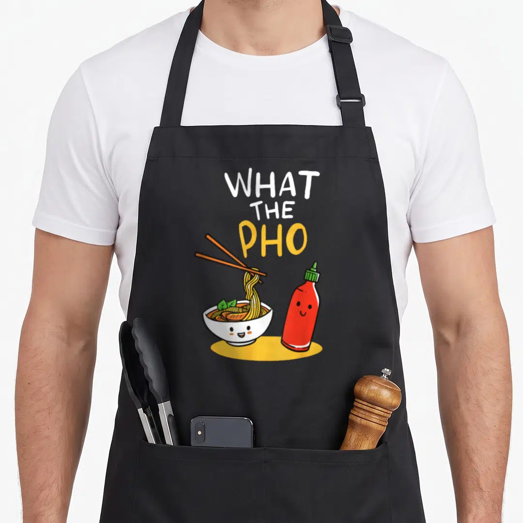 Noodle pho apron - Cartoon bowl of pho with chopsticks and smiling sriracha bottle under stacked text — shown on a black cotton canvas apron against a white background