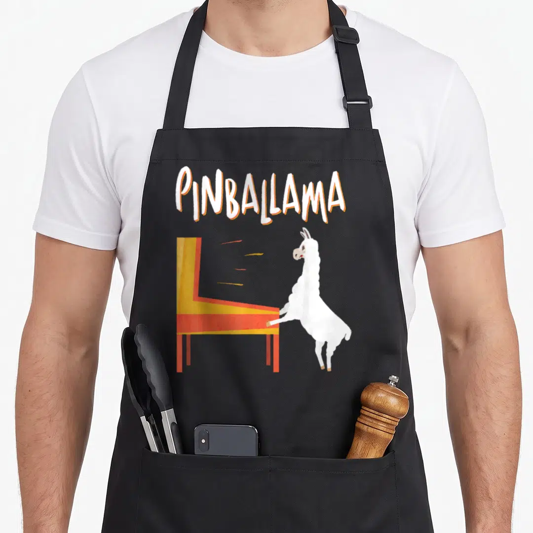Pinballama pinball apron - Arched PINBALLAMA text above a white llama leaning on a yellow and red pinball machine — shown on a black cotton canvas apron against a white background