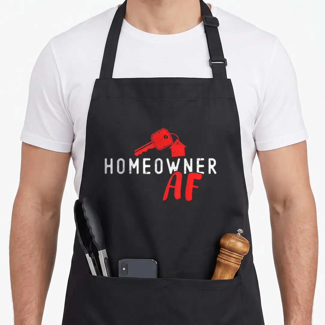 Proud homeowner housewarming apron - Red key with house keychain above white HOMEOWNER and red AF text — shown on a black cotton canvas apron against a white background