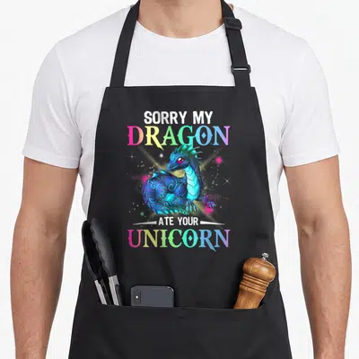 Sorry dragon apron - Blue cartoon dragon with SORRY MY DRAGON ATE YOUR UNICORN text in rainbow gradients — shown on a black cotton canvas apron against a white background