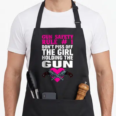 Gun safety apron - Pink and white text reading GUN SAFETY above two crossed revolvers over a pink heart — shown on a black cotton canvas apron against a white background