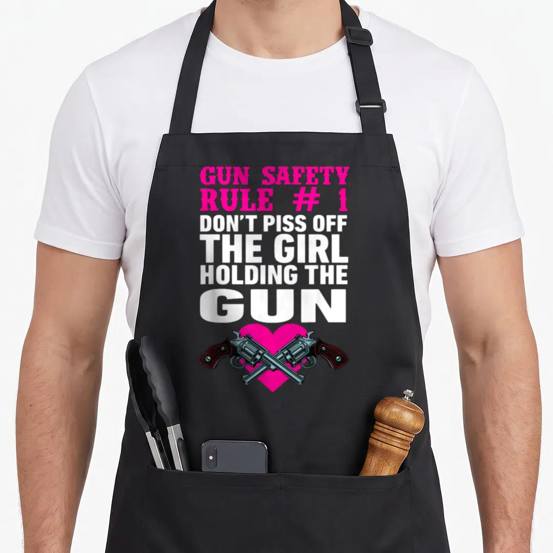 Gun safety apron - Pink and white text reading GUN SAFETY above two crossed revolvers over a pink heart — shown on a black cotton canvas apron against a white background