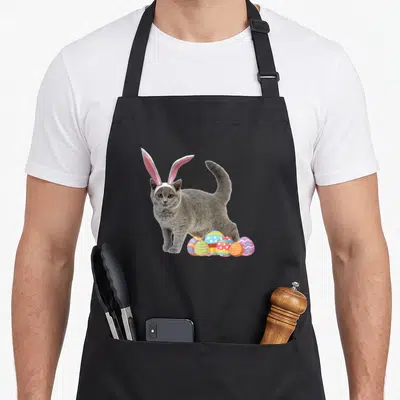 Bunny russian aprons - Grey cat with pink bunny ears beside six patterned easter eggs in blue yellow purple pink — shown on a black cotton canvas apron against a white background