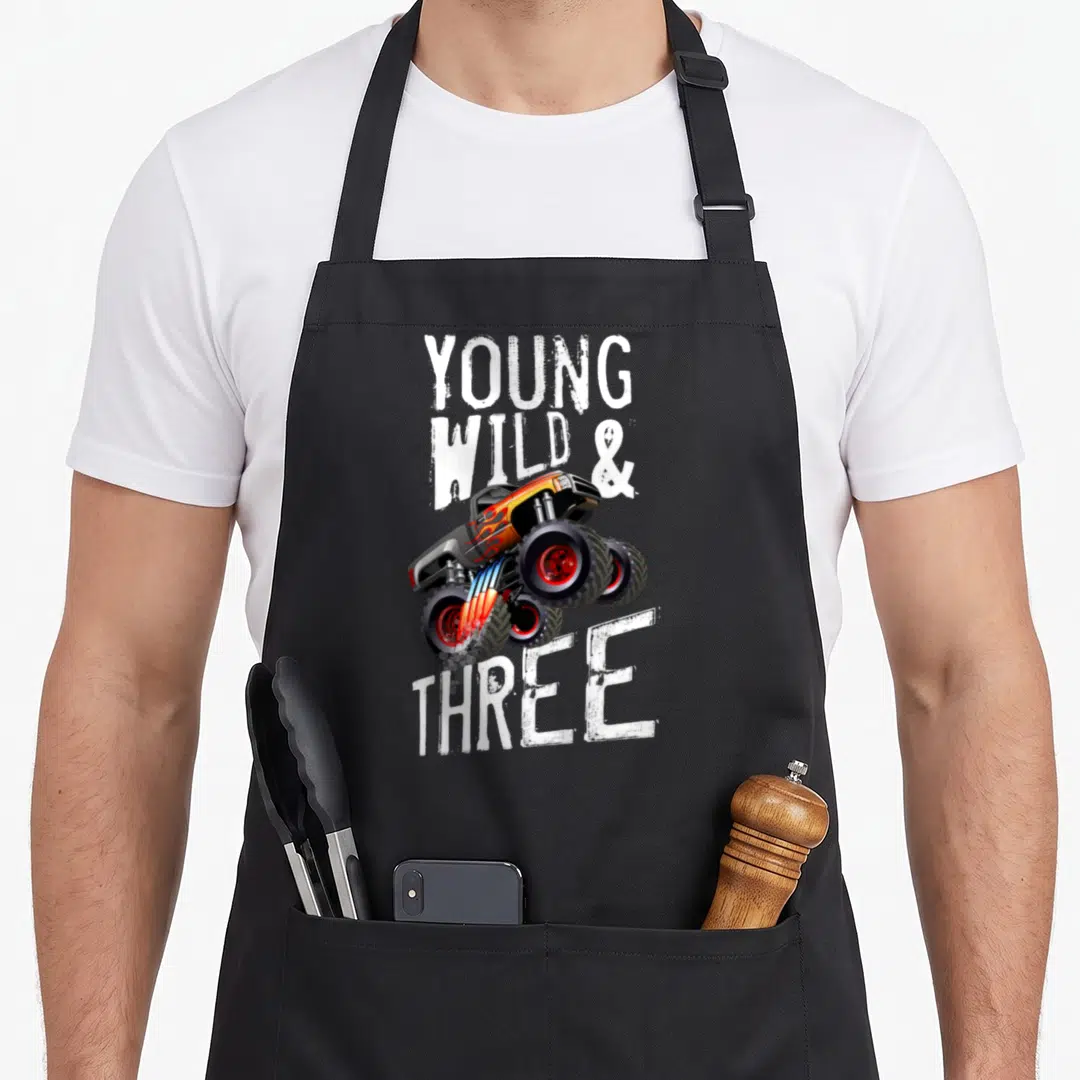 Monster rule truck apron - Cartoon monster truck with YOUNG WILD & THREE text in orange and black — shown on a black cotton canvas apron against a white background
