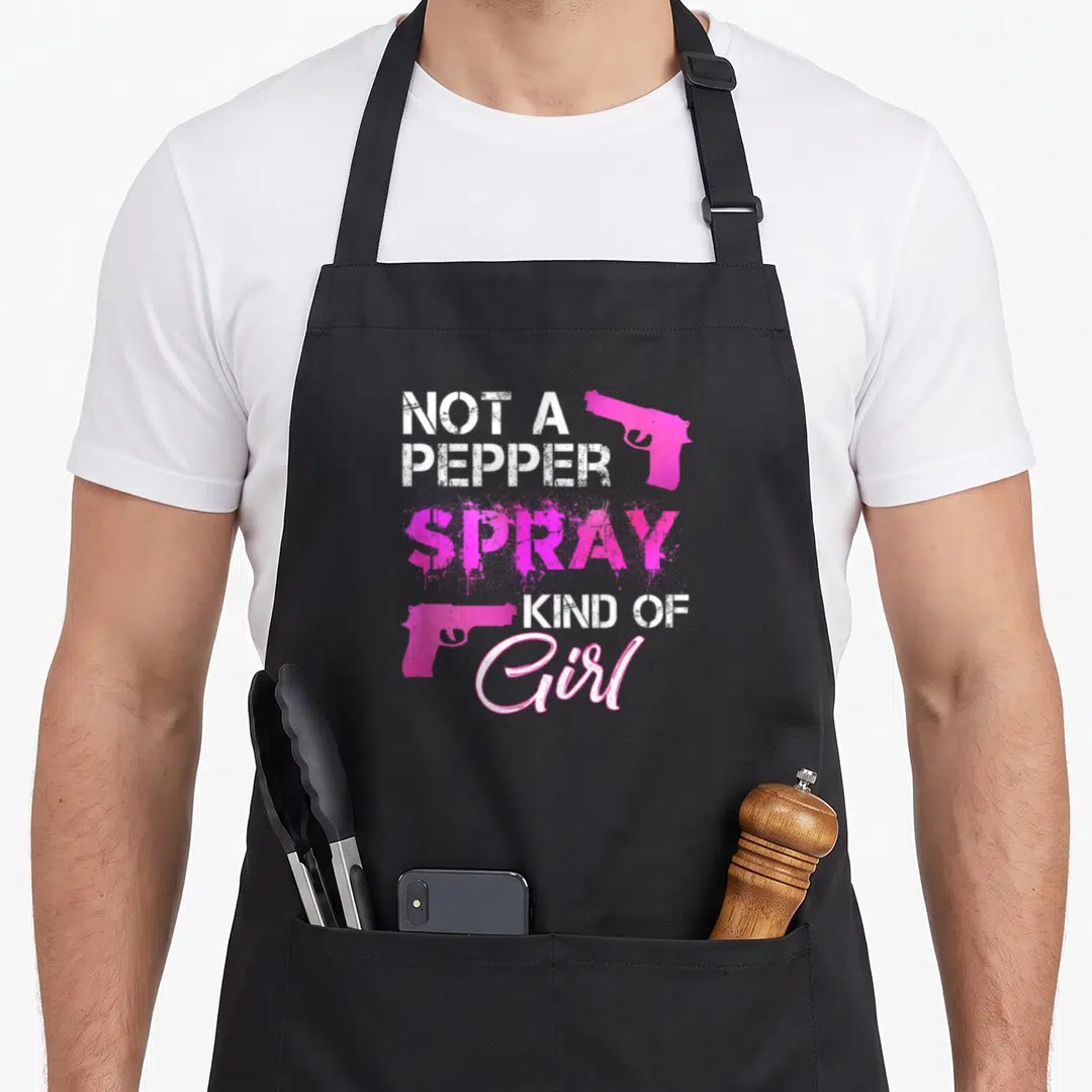 Pepper gun apron - Distressed text Not A Pepper Spray Kind Of Girl with two pink handguns — shown on a black cotton canvas apron against a white background
