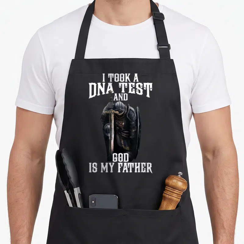 Dna test kitchen apron - Realistic knight holding vertical sword with white text I took a DNA test and God is my father — shown on a black cotton canvas apron against a white background