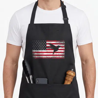 Hummingbird 4th of july apron - Grey hummingbird silhouette over distressed red white American flag — shown on a black cotton canvas apron against a white background