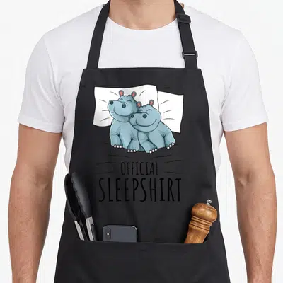 Hippopotamus hippo mom apron - Two blue-grey hippos lying on white pillow with OFFICIAL SLEEPSHIRT text below — shown on a black cotton canvas apron against a white background
