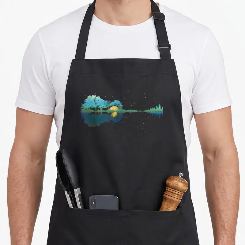 Guitar sky apron - Symmetrical landscape with blue trees, yellow sun, and starry reflection in water — shown on a black cotton canvas apron against a white background