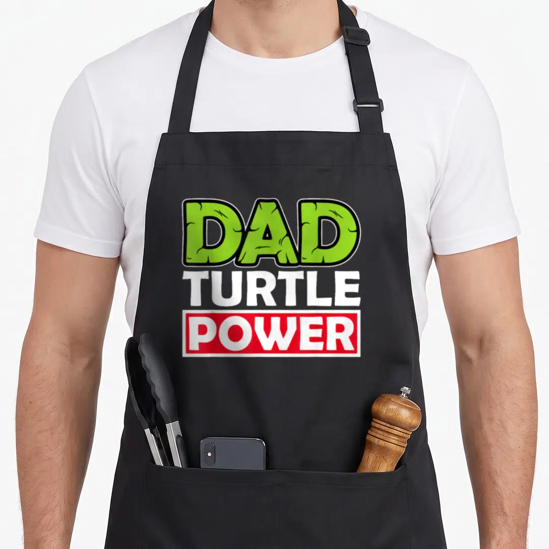 Power father day apron - DAD TURTLE POWER text in green, white, and red stacked typography — shown on a black cotton canvas apron against a white background