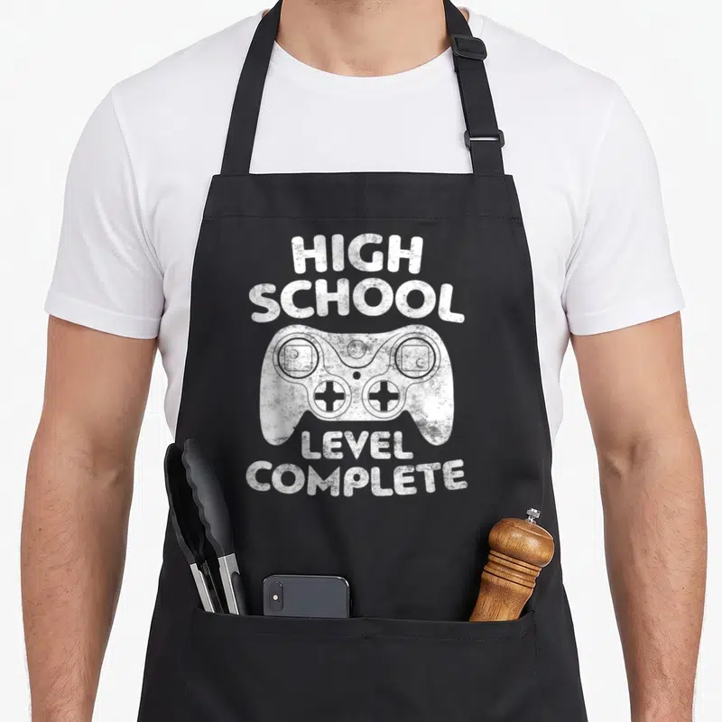 Level complete school graduation apron - White distressed game controller with arched HIGH SCHOOL and stacked LEVEL — shown on a black cotton canvas apron against a white background