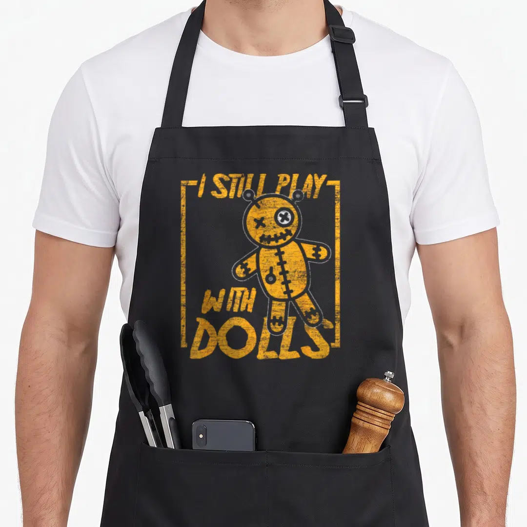 Karma halloween apron - Distressed orange voodoo doll graphic with text I STILL PLAY WITH DOLLS enclosed — shown on a black cotton canvas apron against a white background