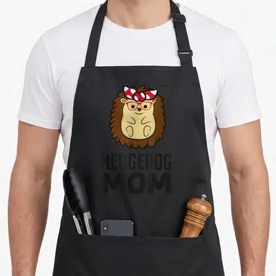 Hedgehog mommy apron - Cartoon brown hedgehog with red glasses and bow above stacked black text HEDGEHOG MOM — shown on a black cotton canvas apron against a white background