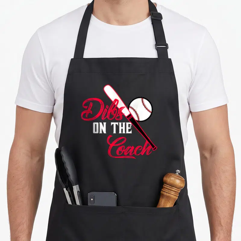 Dib coach baseball wife apron - Red and white text Dibs on the Coach with diagonal baseball bat and ball — shown on a black cotton canvas apron against a white background