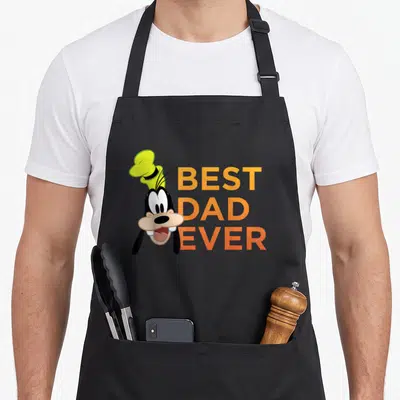 Funny friend father day apron - Goofy head next to orange BEST DAD EVER text on black background — shown on a black cotton canvas apron against a white background