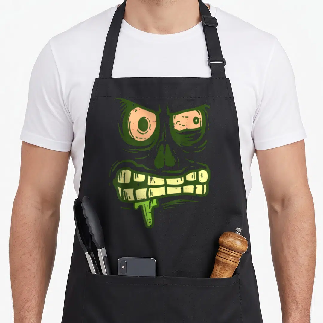 Zombie monster halloween apron - Green cartoon monster face with peach eyes and yellow teeth dripping drool — shown on a black cotton canvas apron against a white background