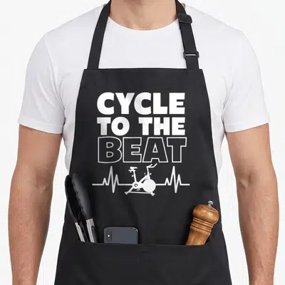 Class spinning apron - White CYCLE TO THE BEAT text above a heartbeat line containing a stationary bike silhouette — shown on a black cotton canvas apron against a white background