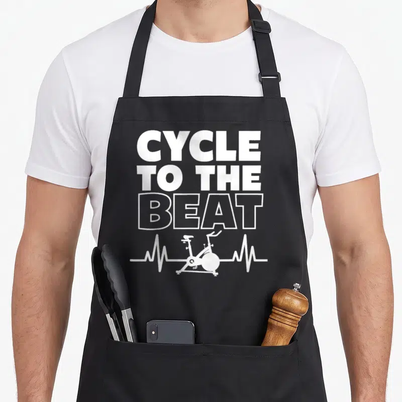 Class spinning apron - White CYCLE TO THE BEAT text above a heartbeat line containing a stationary bike silhouette — shown on a black cotton canvas apron against a white background