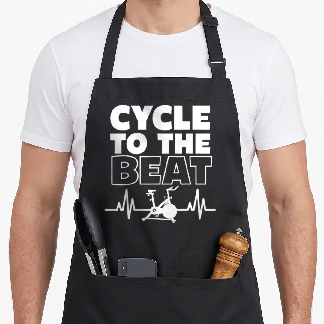 Class spinning apron - White CYCLE TO THE BEAT text above a heartbeat line containing a stationary bike silhouette — shown on a black cotton canvas apron against a white background