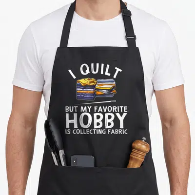 Quilting grandma's aprons - Arched white text above two stacks of folded blue, yellow, and orange fabric with a needle — shown on a black cotton canvas apron against a white background