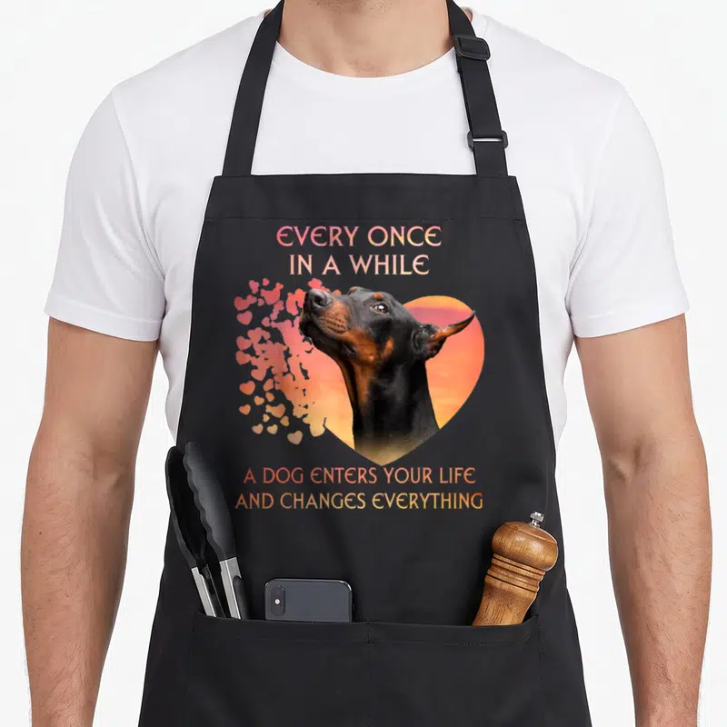 Once dog apron - Doberman head inside orange heart with floating pink hearts and gradient text — shown on a black cotton canvas apron against a white background