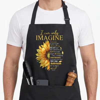 Imagine catholic apron - Yellow watercolor sunflower beside stacked I can only imagine lyrics in white and gold script — shown on a black cotton canvas apron against a white background