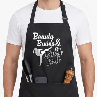 Belt beauty apron - Stacked text Beauty Brains and a Black Belt with kicking martial artist silhouette — shown on a black cotton canvas apron against a white background