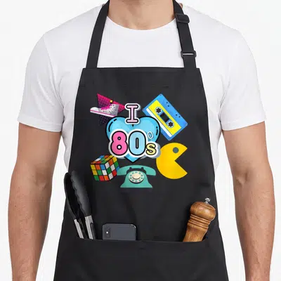 Nostalgic 80s apron - Blue heart graphic with I 80s text surrounded by retro cassette, phone, and game icons in bright — shown on a black cotton canvas apron against a white background