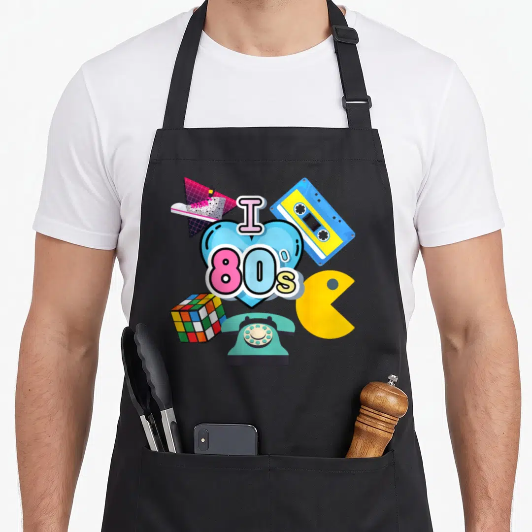 Nostalgic 80s apron - Blue heart graphic with I 80s text surrounded by retro cassette, phone, and game icons in bright — shown on a black cotton canvas apron against a white background