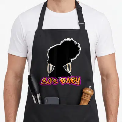 Version 80s apron - Black woman silhouette with gold hoop earrings and curved 80's Baby text in orange and pink — shown on a black cotton canvas apron against a white background