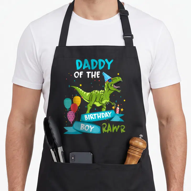 Daddy t-rex apron - Green T-Rex with party hat sunglasses and Daddy of the Birthday Boy text — shown on a black cotton canvas apron against a white background