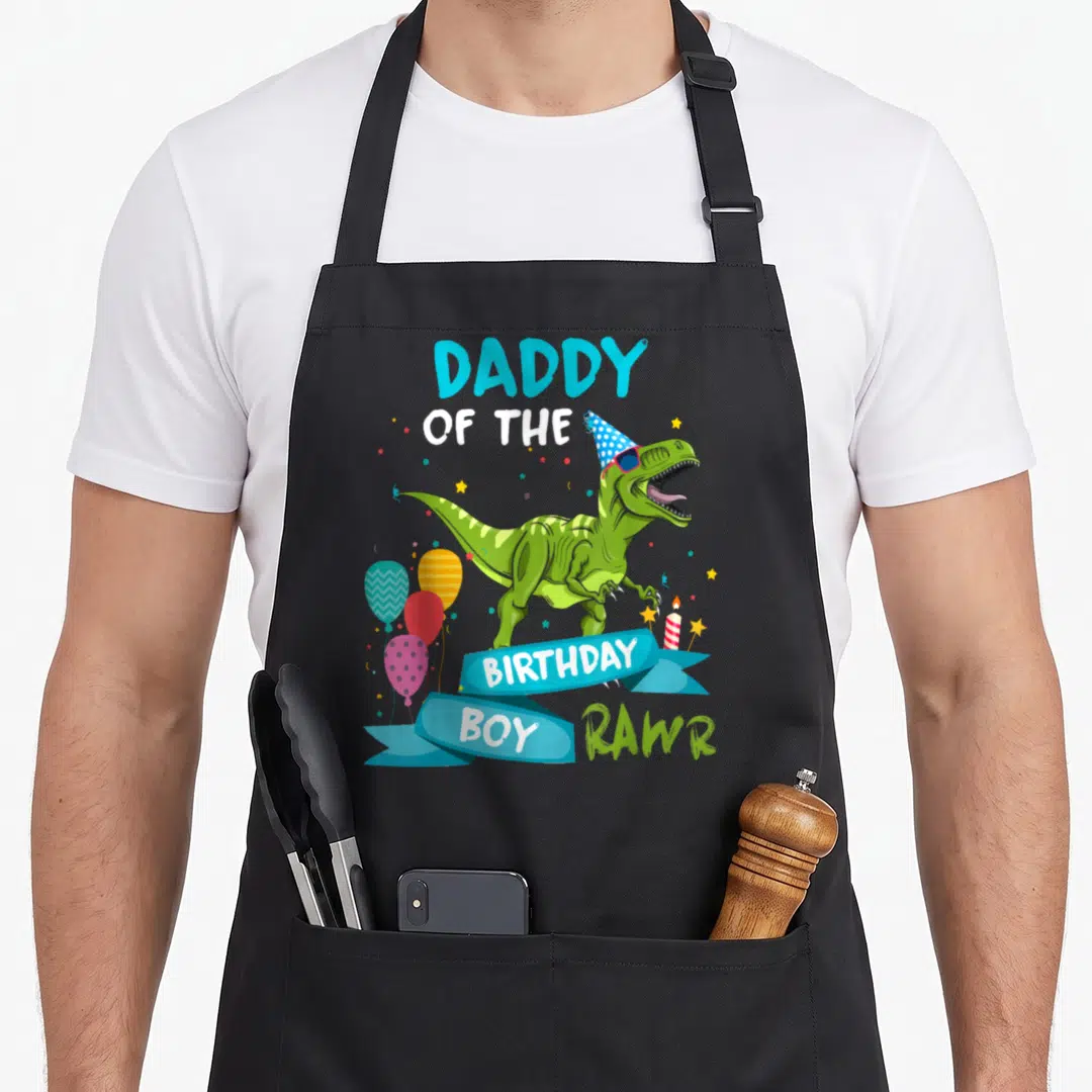 Daddy t-rex apron - Green T-Rex with party hat sunglasses and Daddy of the Birthday Boy text — shown on a black cotton canvas apron against a white background
