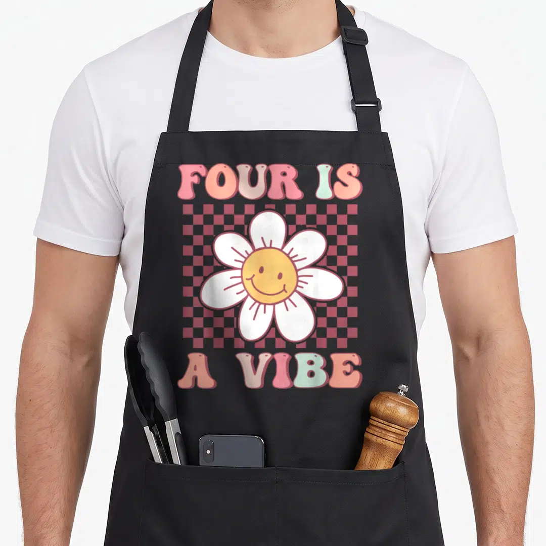 Four daisy apron - Smiling daisy on red pink checkerboard with Four Is A Vibe text — shown on a black cotton canvas apron against a white background