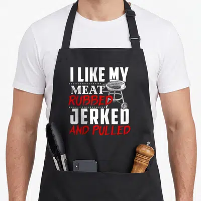 Rubbed meat aprons - Stacked text I LIKE MY MEAT RUBBED JERKED AND PULLED with line-art grill in white and red — shown on a black cotton canvas apron against a white background