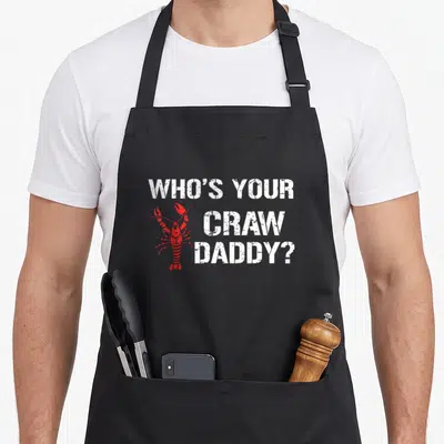 Craw cajun aprons - Red crawfish graphic beside white stacked text WHO'S YOUR CRAW DADDY? in distressed font — shown on a black cotton canvas apron against a white background