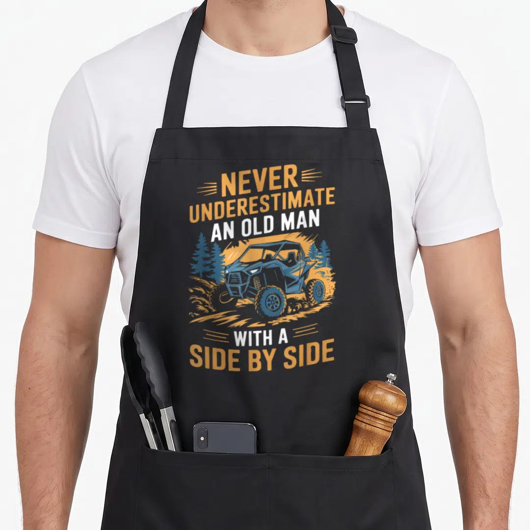 Funny underestimate utv apron - Orange and white text surrounding a blue cartoon side by side UTV kicking up dirt among — shown on a black cotton canvas apron against a white background