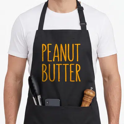 Costume butter apron - Orange distressed PEANUT BUTTER text stacked on grey background — shown on a black cotton canvas apron against a white background