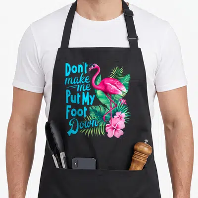 Cute foot pink flamingo apron - Pink flamingo wearing blue glasses standing among green palm leaves next to stacked — shown on a black cotton canvas apron against a white background