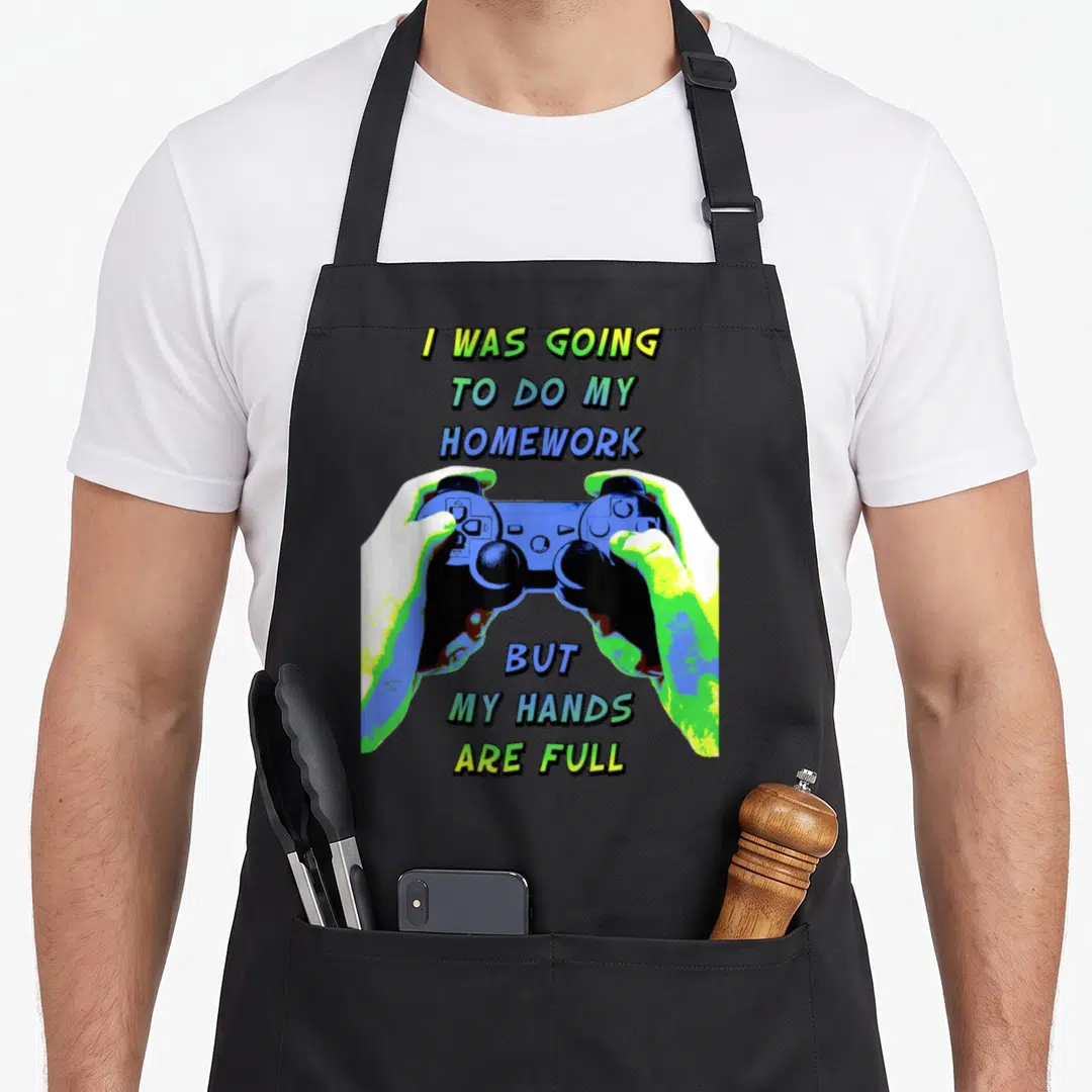 Homework mathware apron - Cartoon hands holding blue game controller with split text about homework and full hands — shown on a black cotton canvas apron against a white background