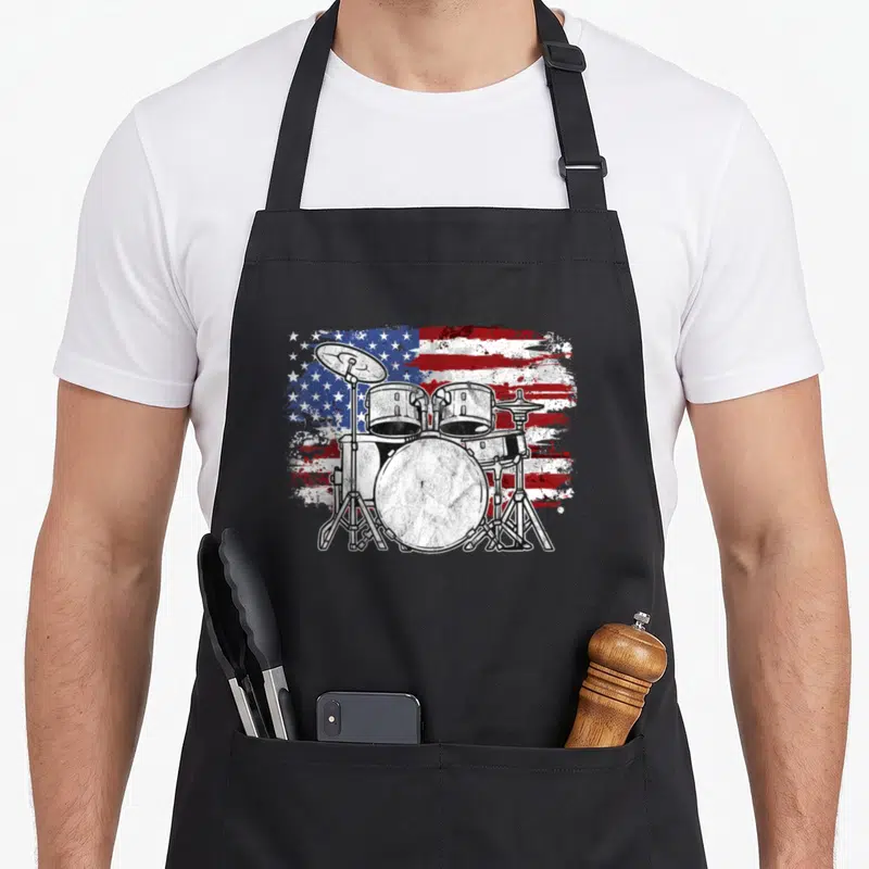 Drummer 4th of july apron - Distressed drum kit illustration overlaid on a grunge style American flag pattern — shown on a black cotton canvas apron against a white background