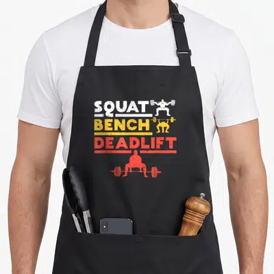 Deadlift bench apron - SQUAT BENCH DEADLIFT text with white yellow red lifter silhouettes — shown on a black cotton canvas apron against a white background
