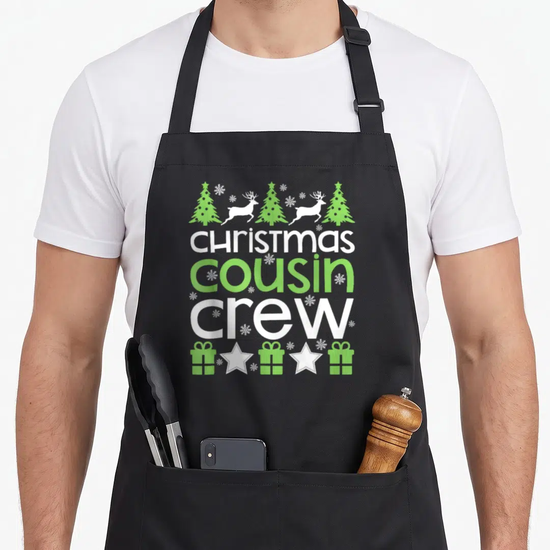 Cousin family christmas aprons - Stacked CHRISTmas cousin CREW text with reindeer, trees, gifts, and snowflakes — shown on a black cotton canvas apron against a white background