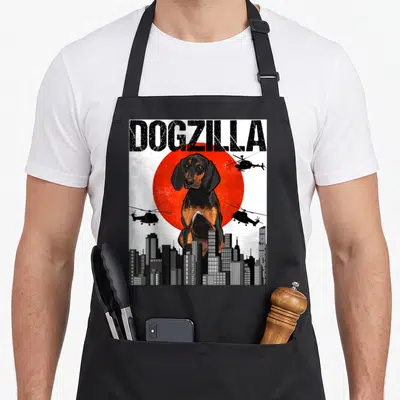 Japanese crazy dog apron - DOGZILLA text above black tan dog, red sun, helicopters, grey skyline — shown on a black cotton canvas apron against a white background