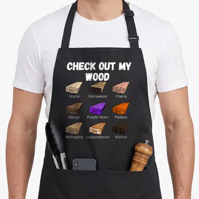 Check woodworking apron - Nine cartoon wood plank stacks labeled Maple, Zebrawood, Cherry, Wenge, Purple Heart, Padauk — shown on a black cotton canvas apron against a white background