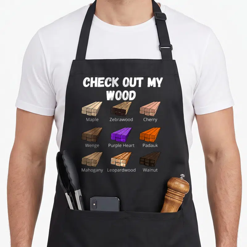Check woodworking apron - Nine cartoon wood plank stacks labeled Maple, Zebrawood, Cherry, Wenge, Purple Heart, Padauk — shown on a black cotton canvas apron against a white background