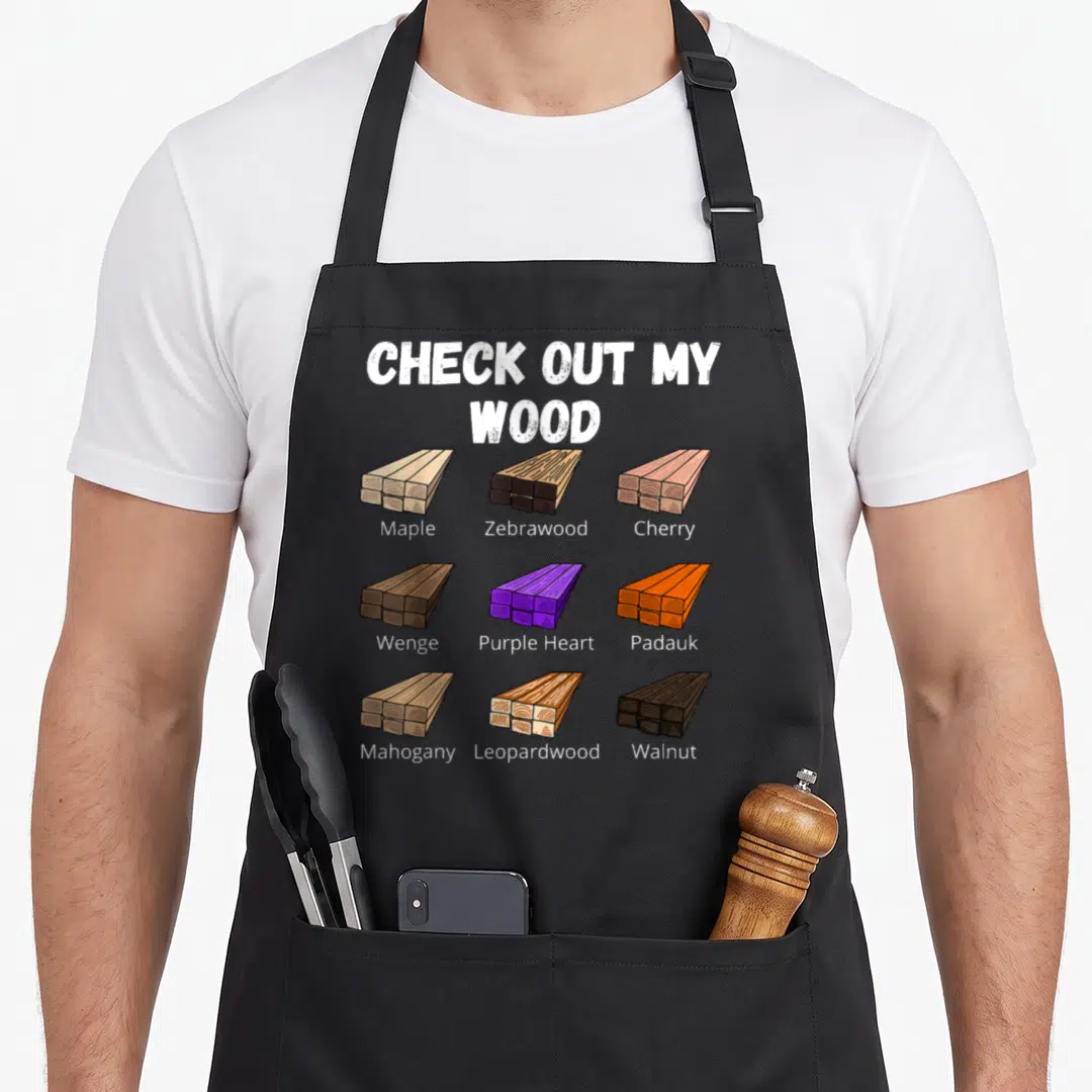 Check woodworking apron - Nine cartoon wood plank stacks labeled Maple, Zebrawood, Cherry, Wenge, Purple Heart, Padauk — shown on a black cotton canvas apron against a white background